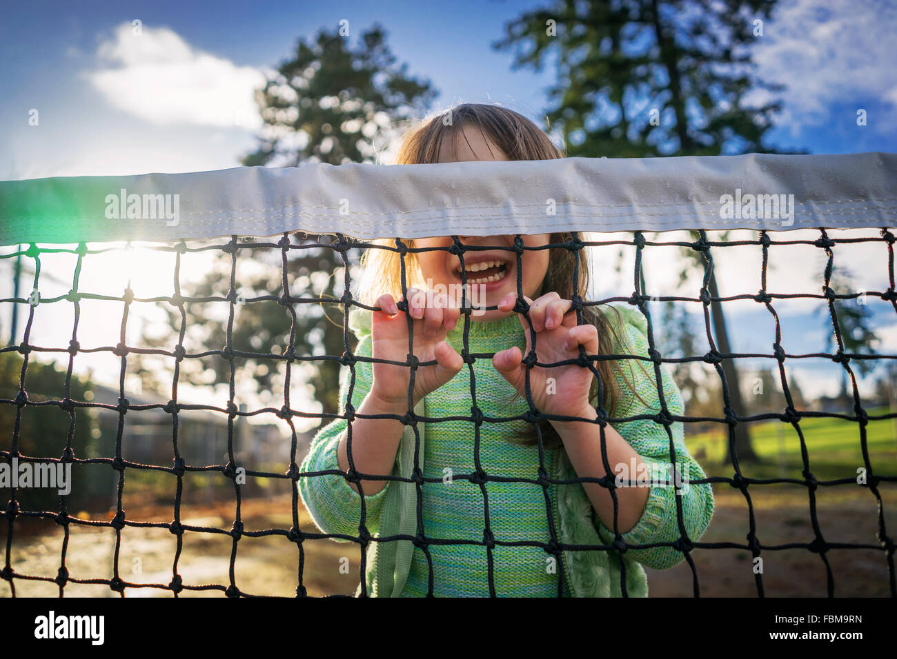 Girl hiding behind tennis net laughing Stock Photo