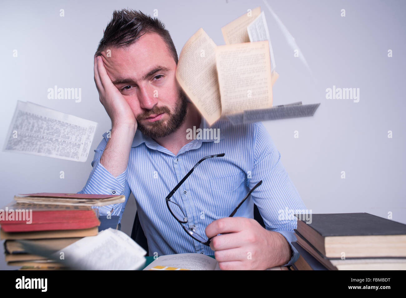 Frustrated man resting head in hand with books and papers flying around Stock Photo
