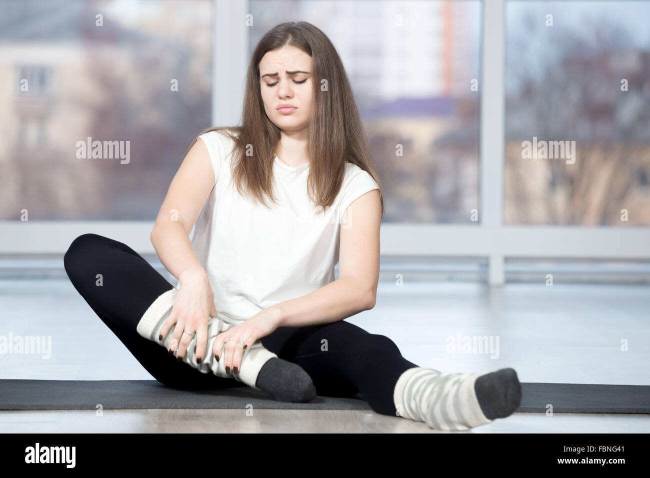 Portrait of injured unhappy fit young beautiful woman sitting in sports club, touching her ankle after working out in class Stock Photo
