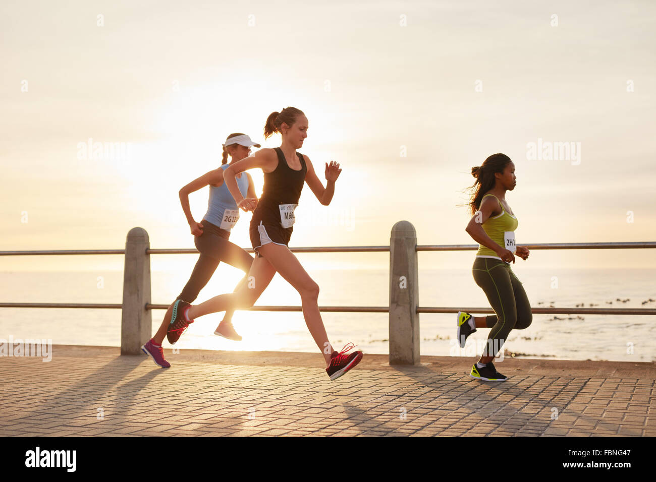 Three young women running on a road by the sea. Group of divers runners training on seaside promenade during sunset. Stock Photo