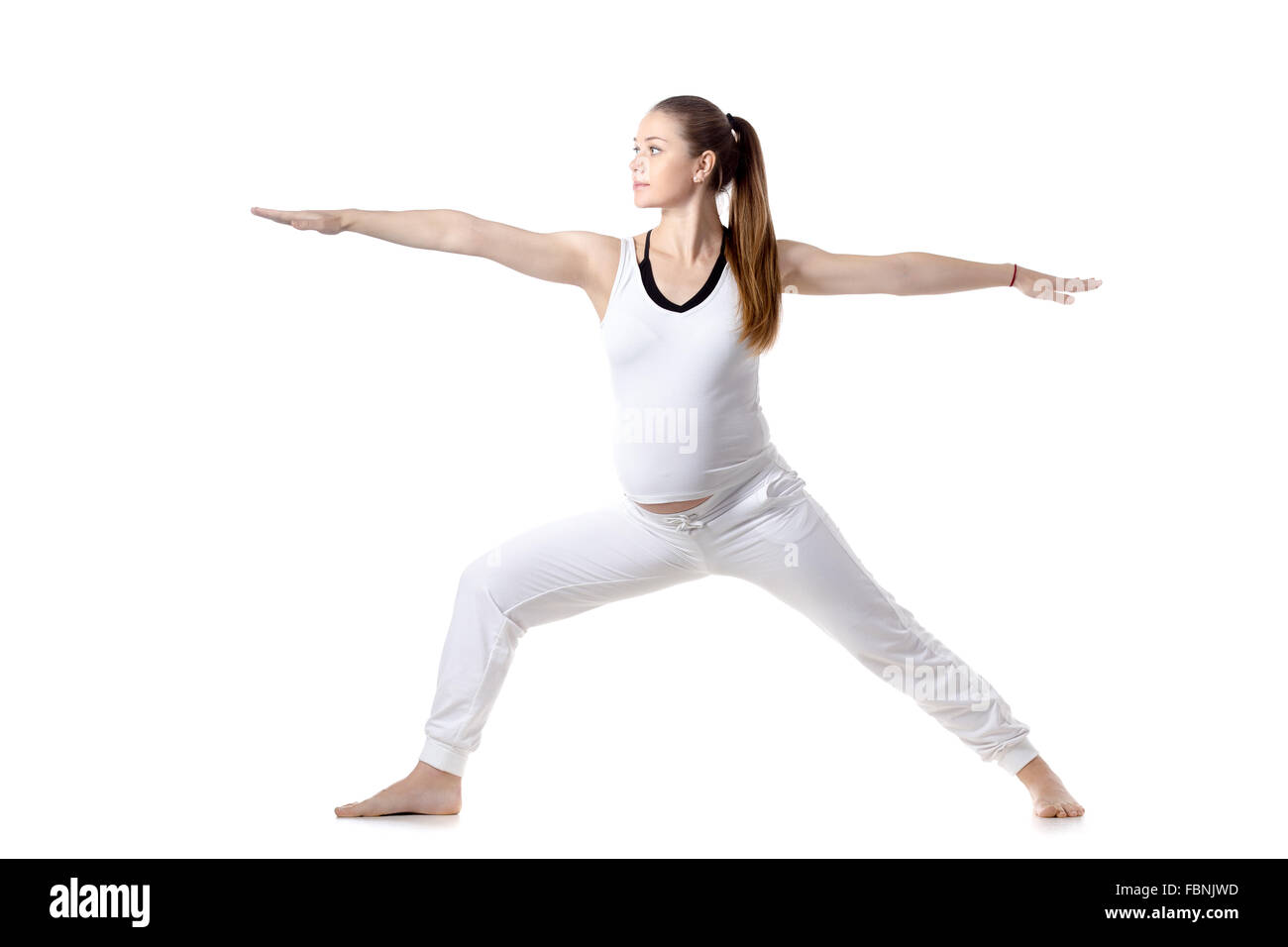 Full length portrait of young pregnant fitness model in white sportswear doing yoga or pilates training, standing in Warrior II Stock Photo