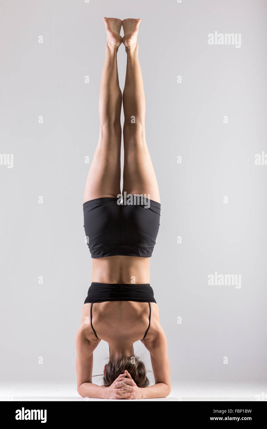 Sporty girl doing Supported Headstand, yoga asana Sirsasana, Shirshasana, Sirshasana, Headstand on grey background, low key shot Stock Photo