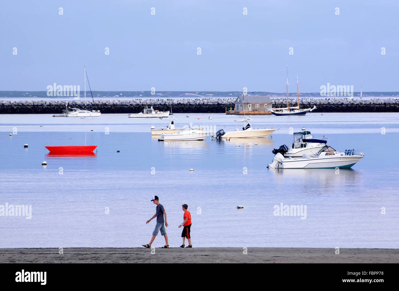 Two young boys walking past a moored red dory, motorboats, and sailboats at Provincetown Harbor at sunset. Stock Photo