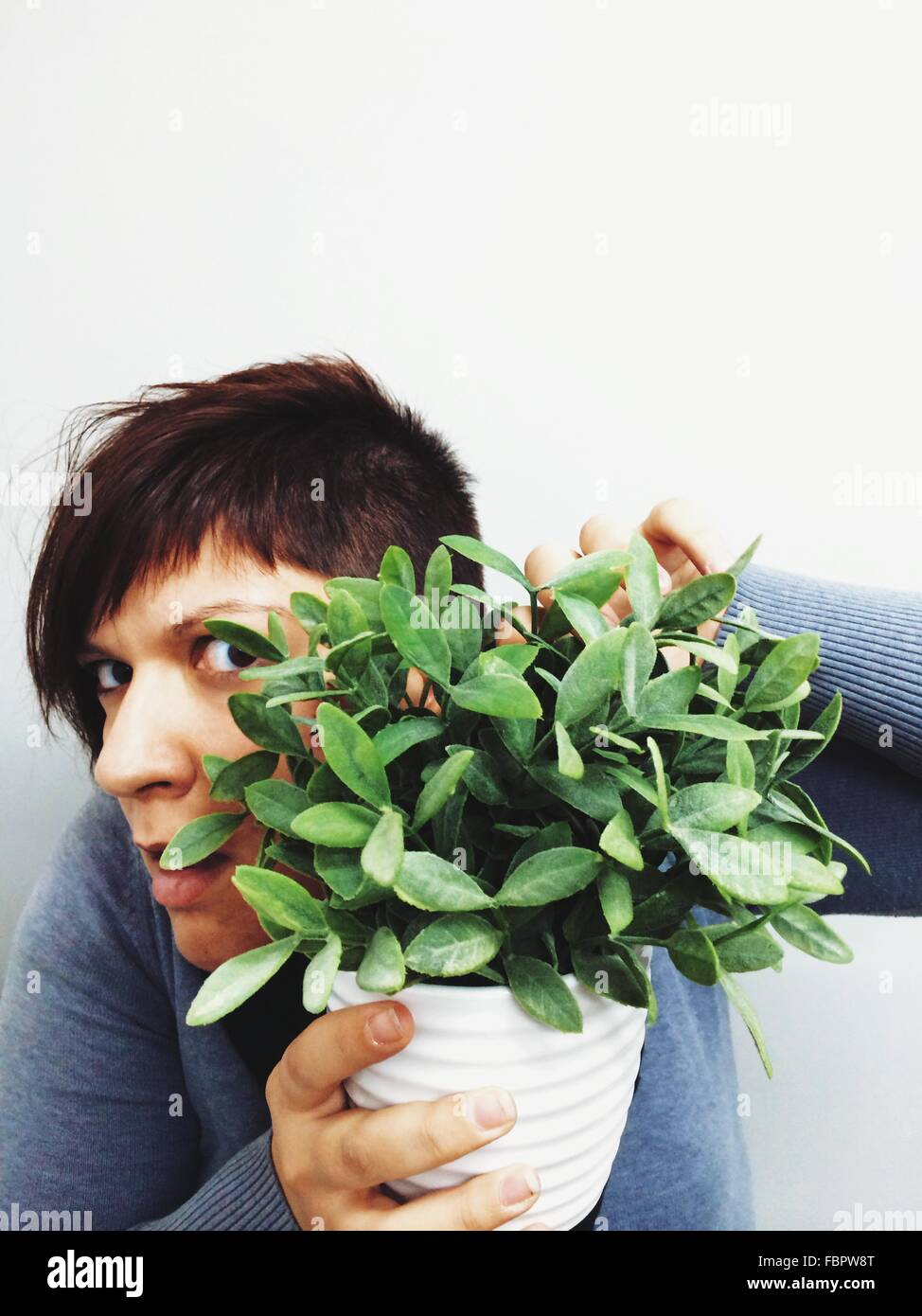 Portrait Of Suspicious Young Woman Hiding Behind Potted Plant Over White Background Stock Photo
