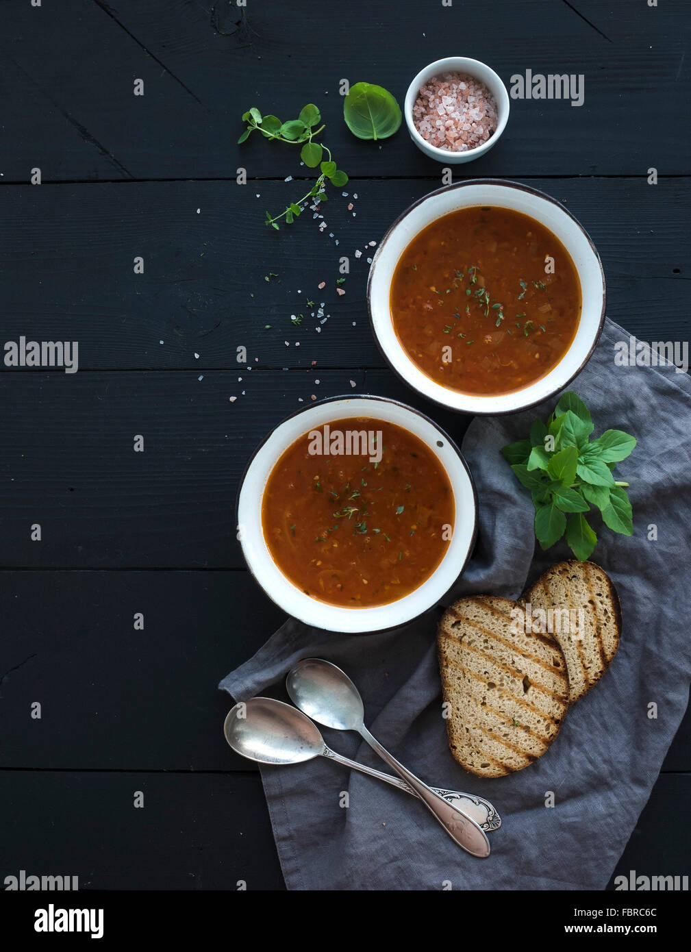 Roasted tomato soup with fresh basil, spices and bread in rustic metal bowls over black background, top view, copy space Stock Photo