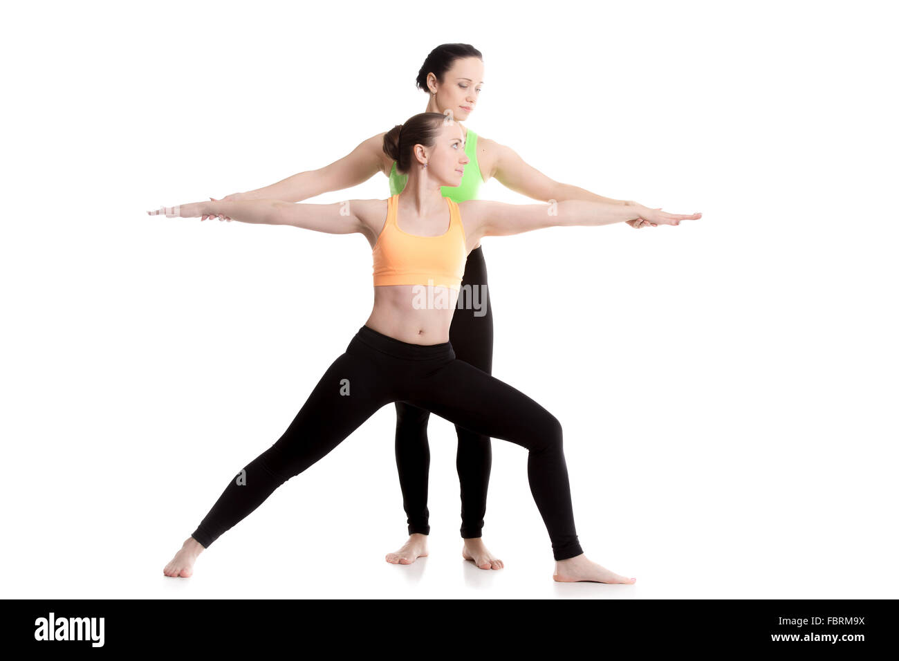 Two beautiful sporty girls practice yoga with partner, coach helps student, doing lunge exercise, Warrior II posture, Virabhadra Stock Photo