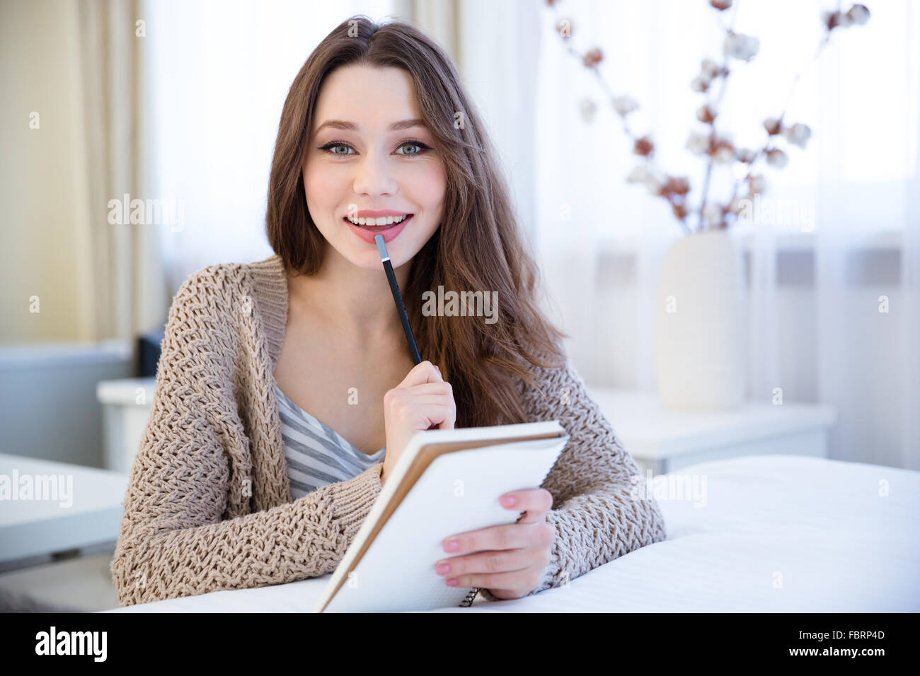 Charming cheerful young woman thinking and writing in notepad sitting ...