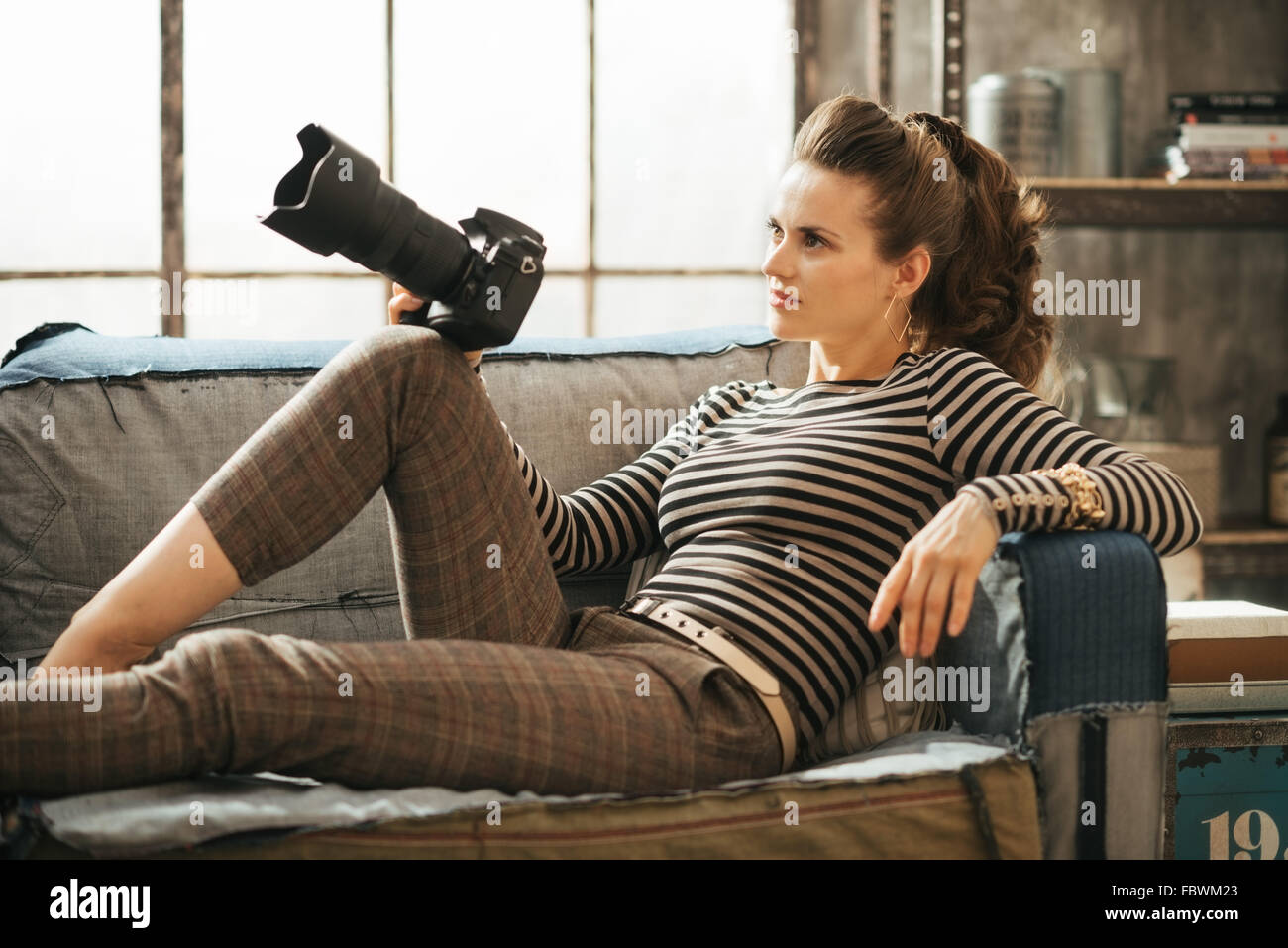 Stylish brown-haired female is sitting on couch and holding dslr photocamera in loft apartment Stock Photo