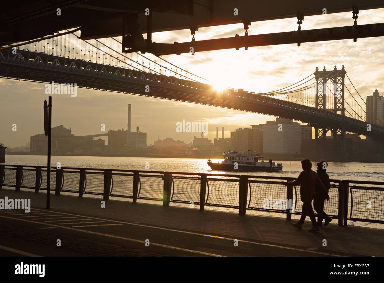 Two women walk north on the East River Esplanade as the sun rises over the Manhattan Bridge New York City Stock Photo