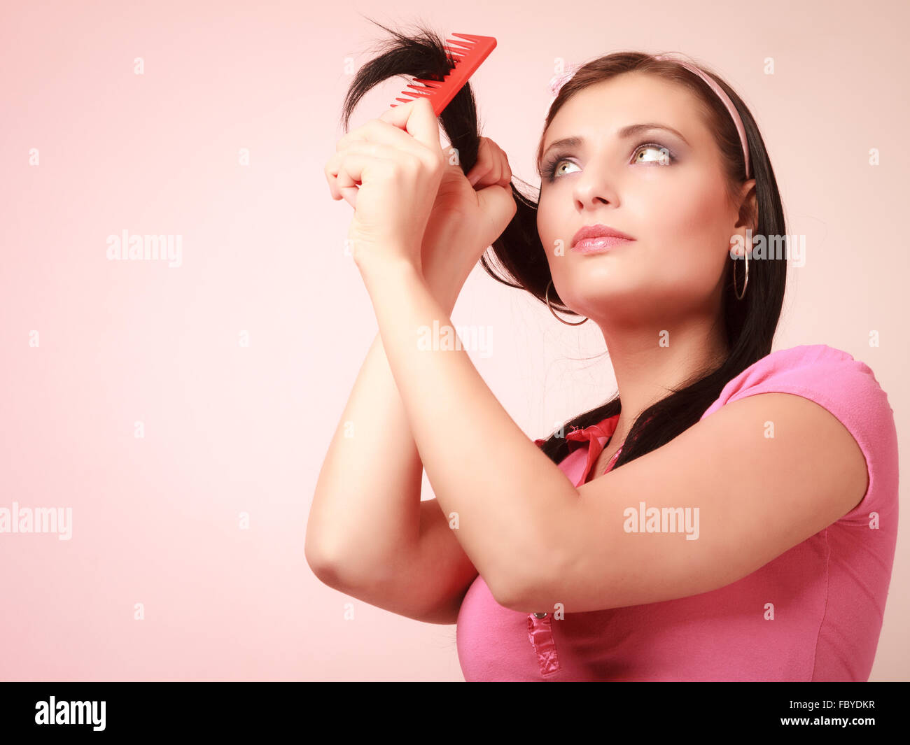 Portrait of childish thoughtful young woman combing her hair and dreaming. Infantile pensive girl with comb on pink. Longing for Stock Photo