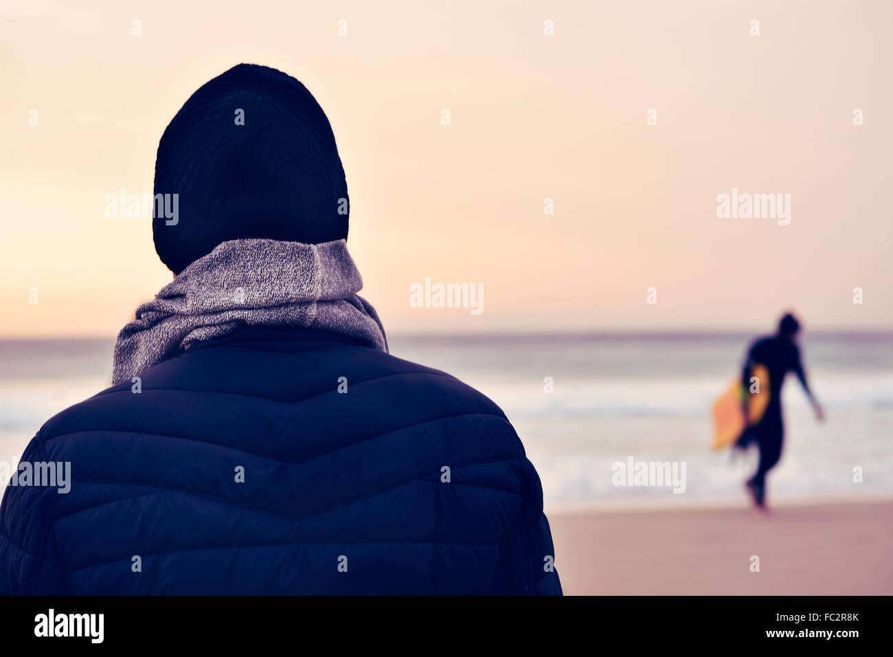 closeup of a young caucasian man seen from behind wearing scarf, a knit cap and a coat in front of the ocean, and a surfer carry Stock Photo