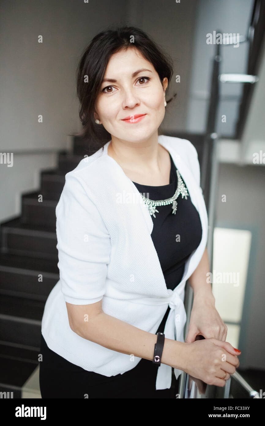 Pretty brunette business woman with kind eyes leaning forward posing on stairs of office building, wearing white jacket over tight black dress. Positive look, candid portrait. Consultations, support. Stock Photo