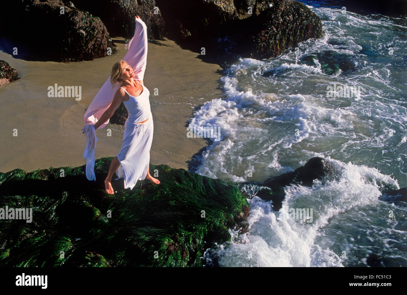 Woman in white dress enjoying life on wave swept rocky shore in California Stock Photo