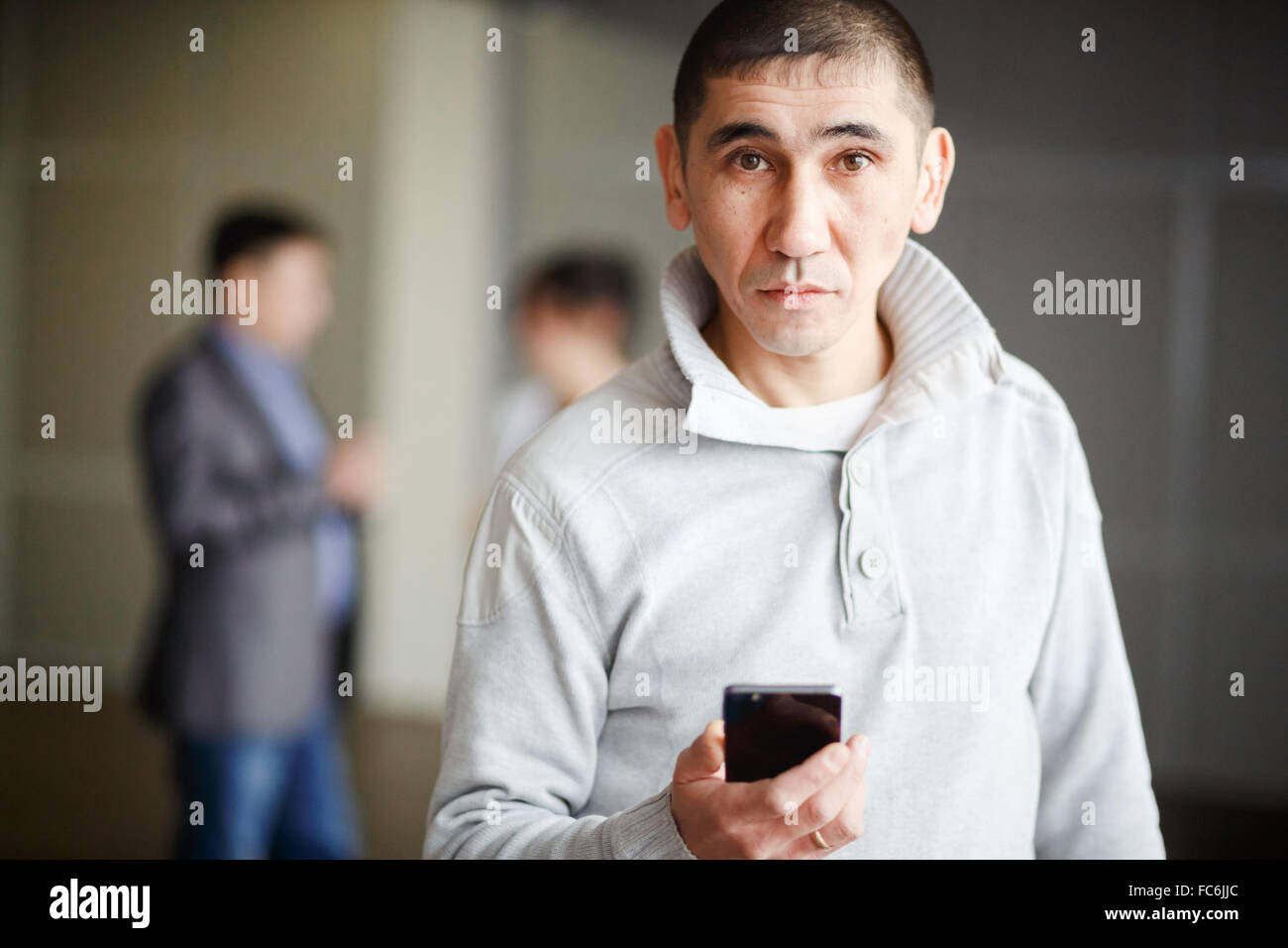 Middle Asian man short haircut with phone in his hand questioning look directly into camera, gets a job. Against background of business men discussing employment. Unemployment, human resources. Stock Photo