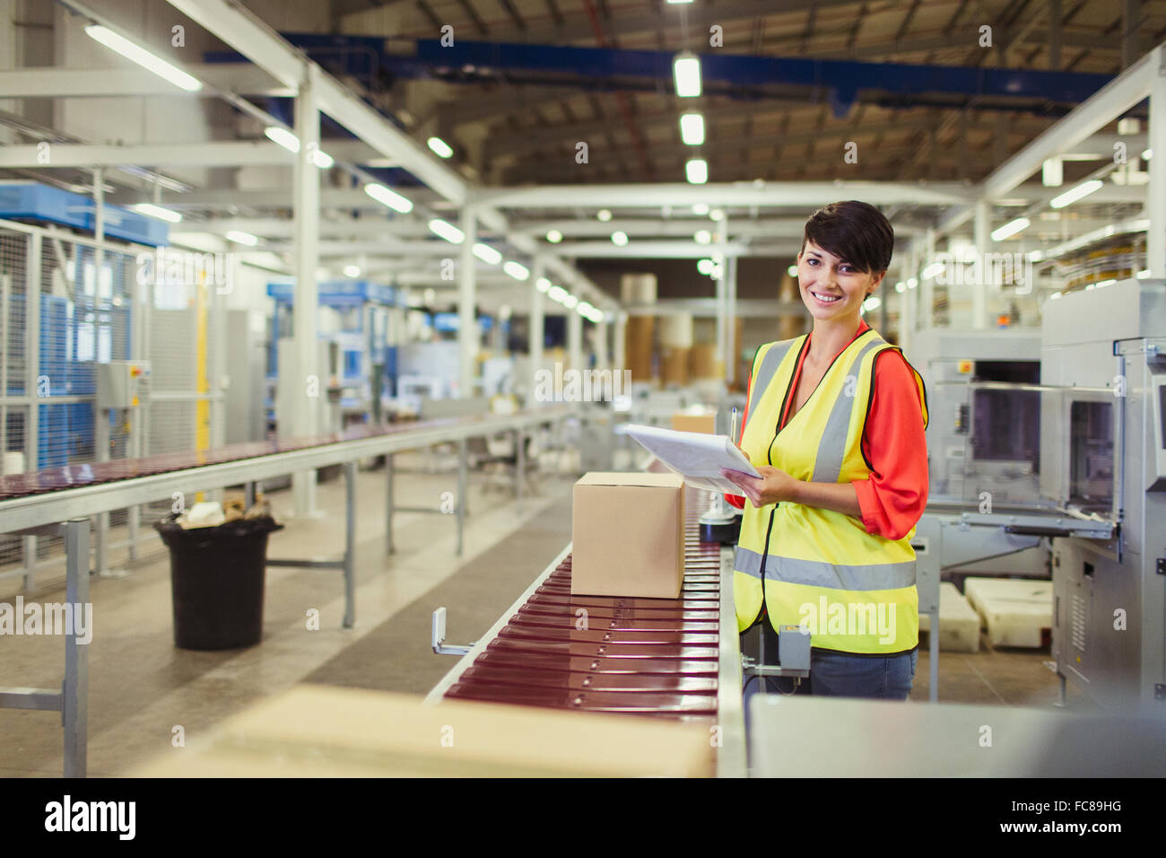 Portrait smiling worker checking cardboard boxes on conveyor belt production line in factory Stock Photo
