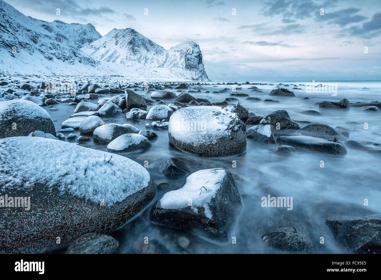 Rocks in the cold sea and snow capped mountains under the blue light of dusk, Unstad, Lofoten Islands, Arctic, Northern Norway Stock Photo