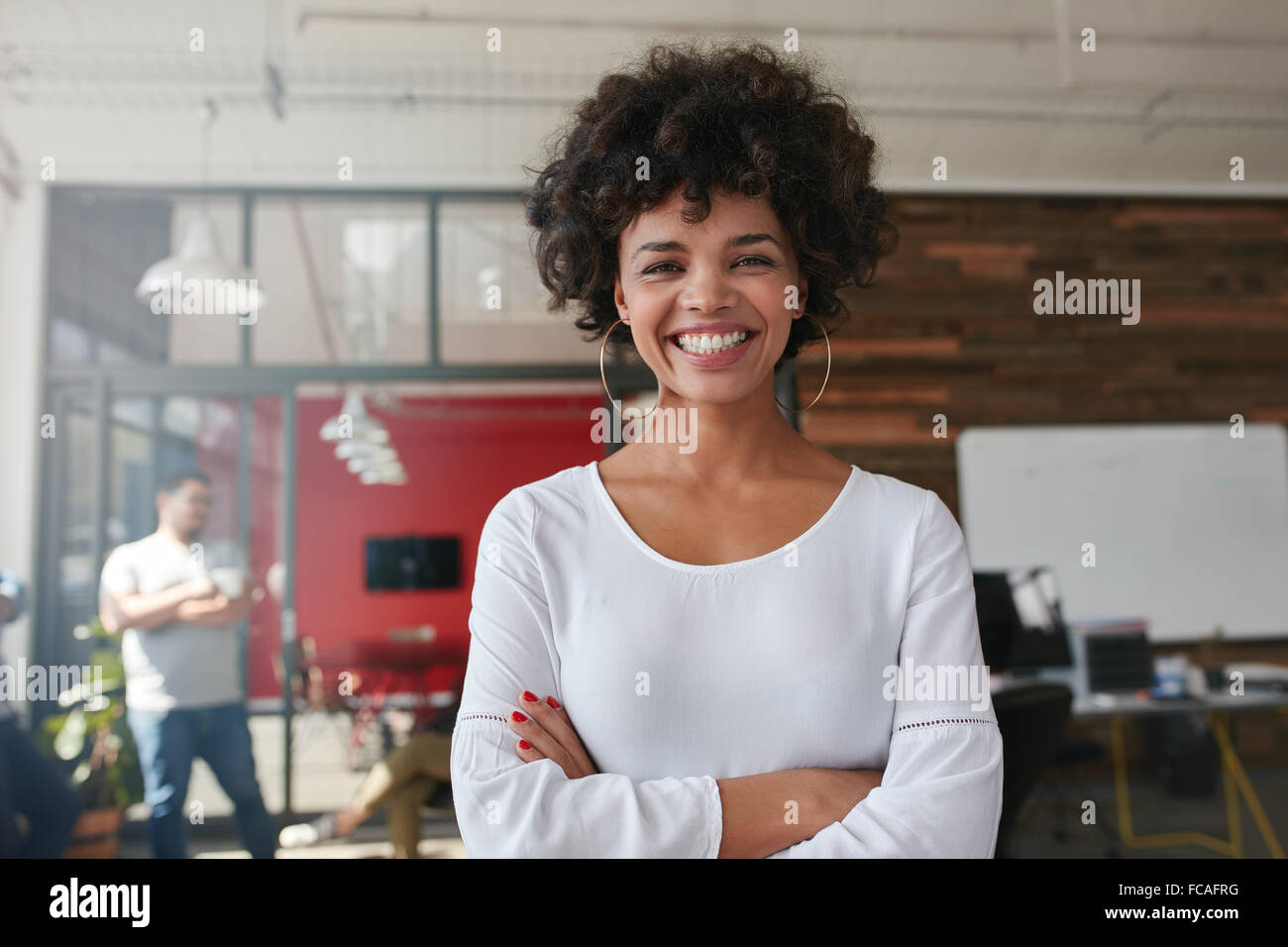 Smiling young woman standing with her arms crossed and looking at camera. She is standing in a modern office with her colleagues Stock Photo