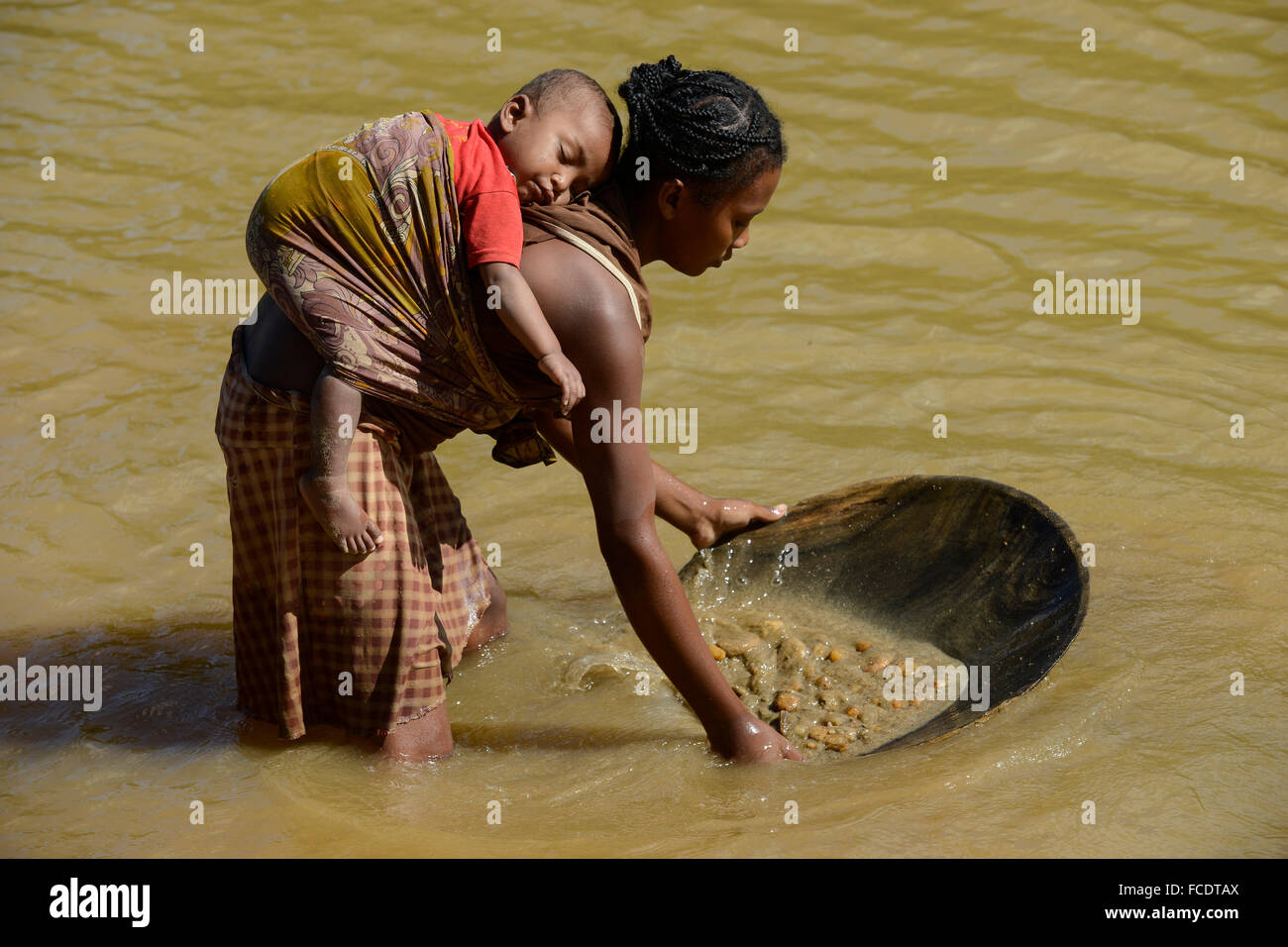 MADAGASCAR, region Manajary, town Vohilava, small scale gold mining, women panning for gold at river ANDRANGARANGA, mother carry her child on the back during the work / MADAGASKAR Mananjary, Vohilava, kleingewerblicher Goldabbau, Frauen waschen Gold am Fluss ANDRANGARANGA, Mutter traegt ihr Kind bei der Arbeit auf dem Ruecken Stock Photo