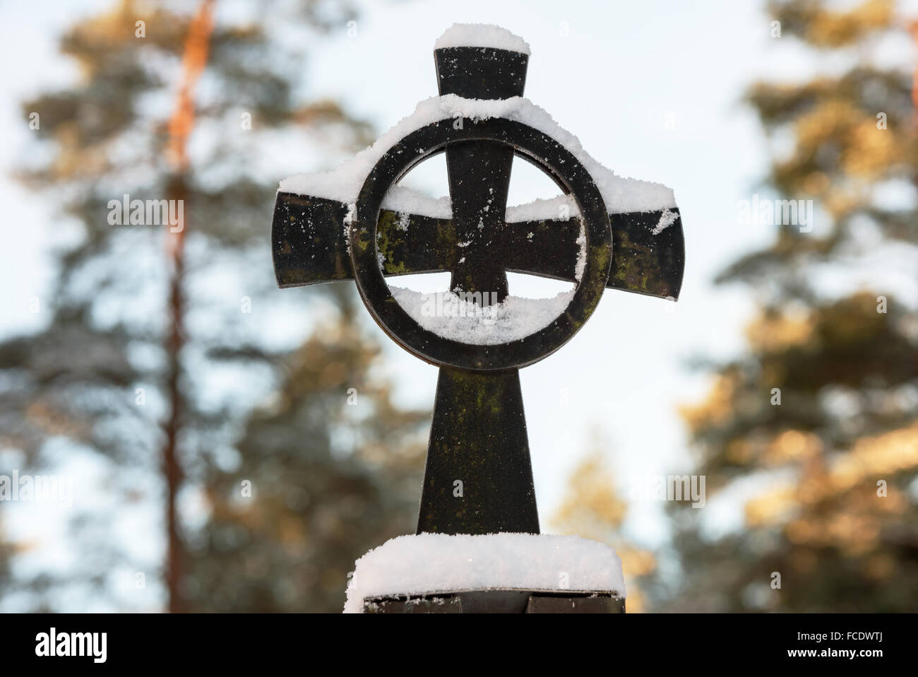 A black Christian iron cross with a circle. Cross is covered with a blanket of snow. Tree in background. Stock Photo