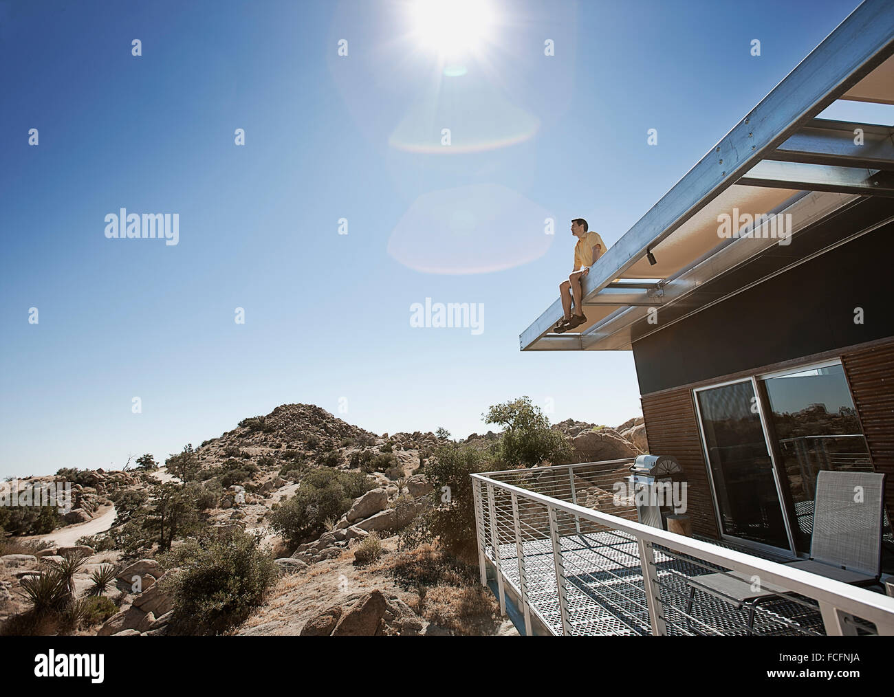 A man sitting on the roof overhang of an eco home in the desert landscape. Stock Photo
