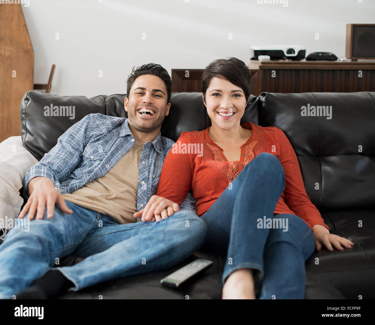 A man and woman sitting on a sofa, side by side, holding hands and watching a screen. Stock Photo