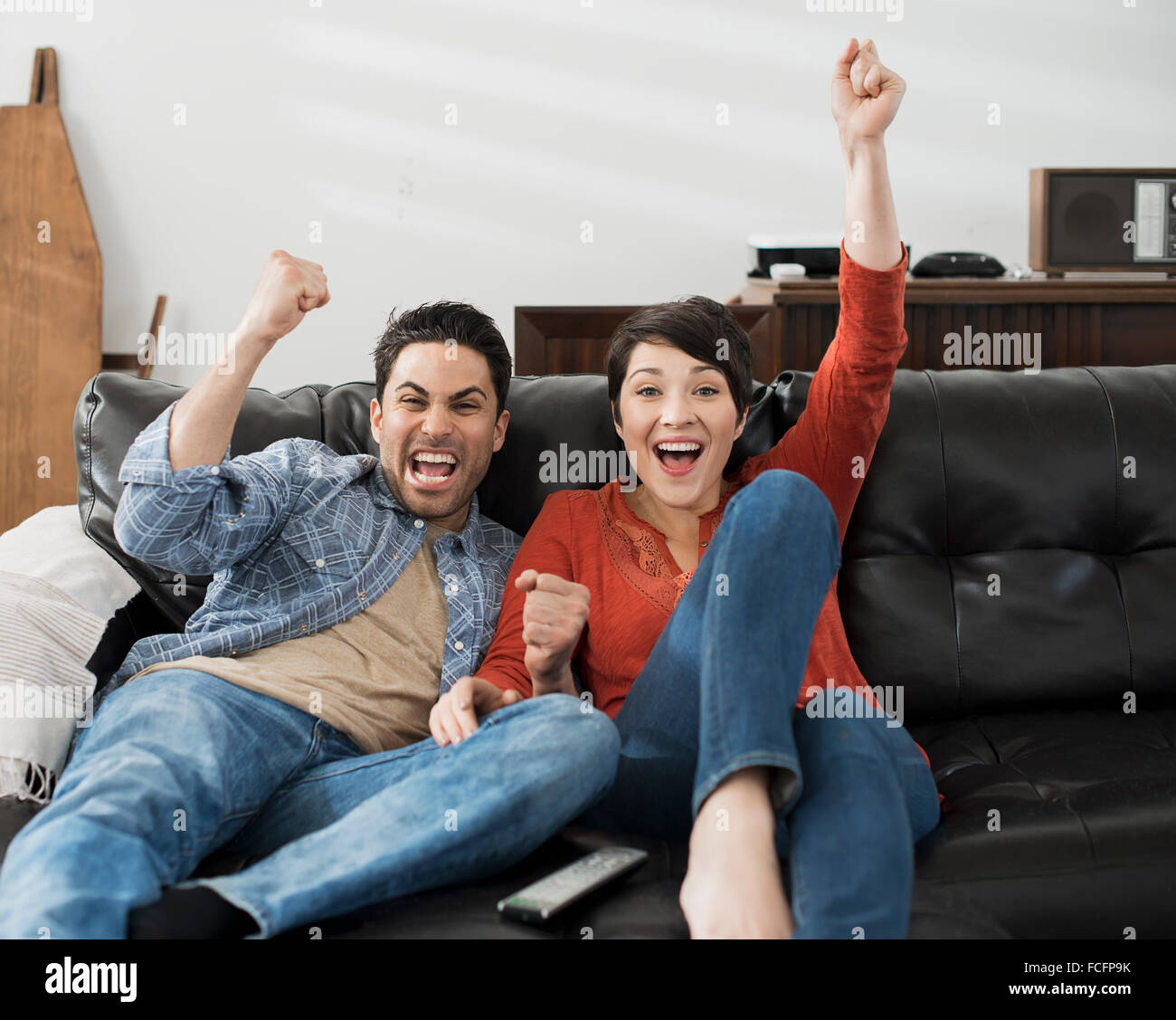 A man and woman sitting on a sofa, celebrating and pumping the air with their fists. Watching sport on the tv. Stock Photo