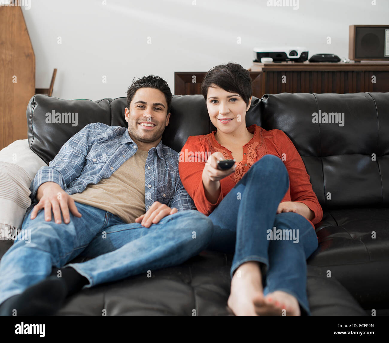 A man and woman sitting on a sofa, side by side, one using the remote control for the tv. Stock Photo