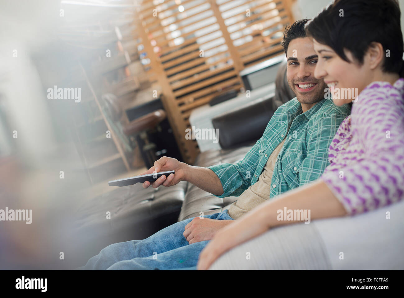 A couple, man and woman sitting on a sofa at home, one holding a remote control device. Stock Photo