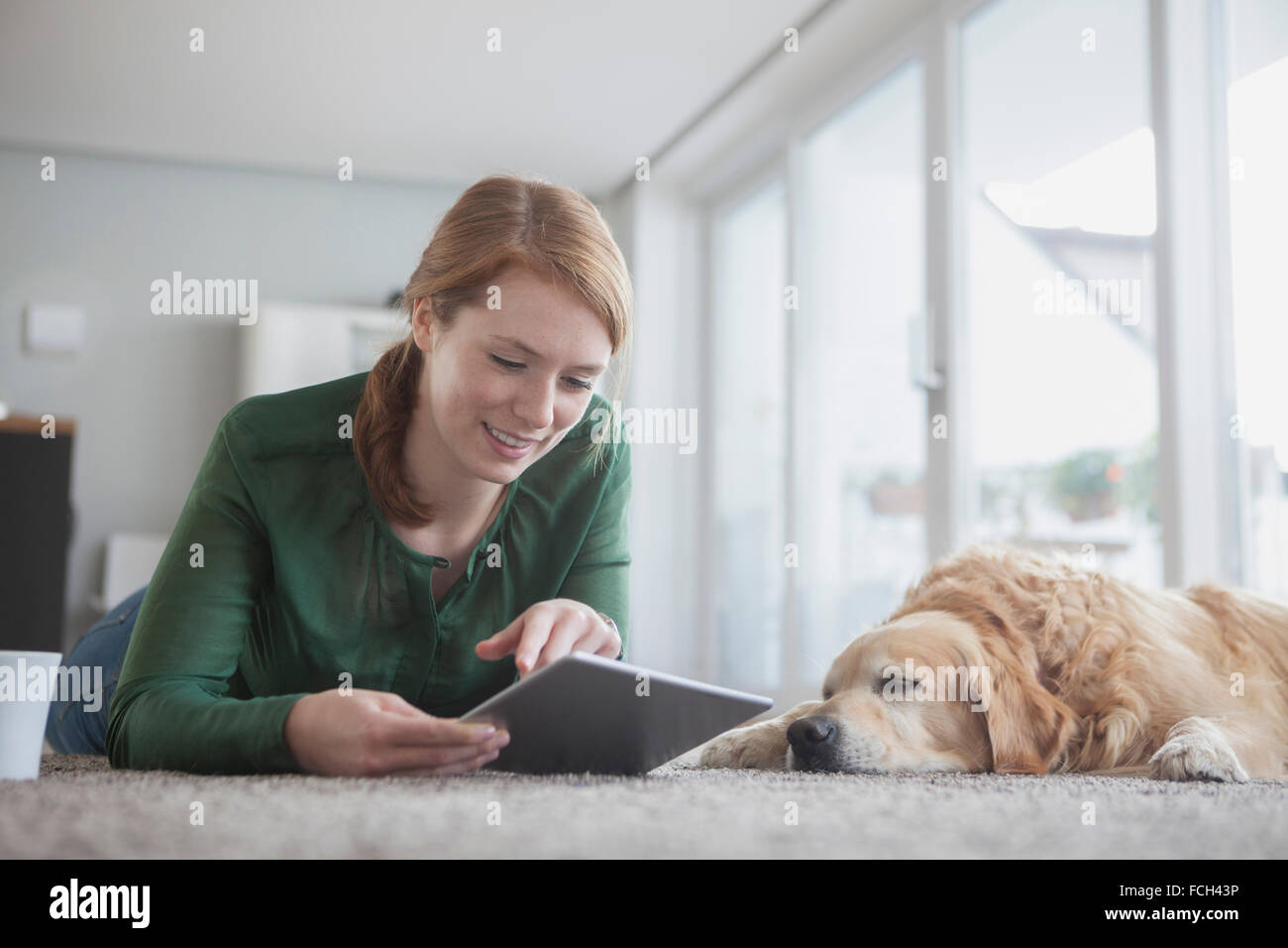 Smiling young woman lying beside her sleeping dog on the carpet using digital tablet Stock Photo