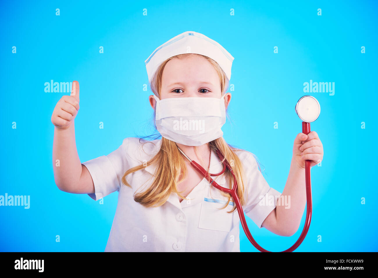 cute girl dressed as a doctor with a stethoscope, examining a patient on a blue background. Stock Photo