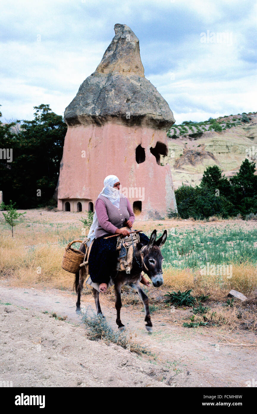 Turkish Peasant Woman Riding a Donkey in Front of a Volcanic Fairy Chimney or Volcanic Hoodoo Converted into a Troglodyte House with Rock-Cut Windows in the Soft Tufa Rock Cappadocia Turkey Stock Photo