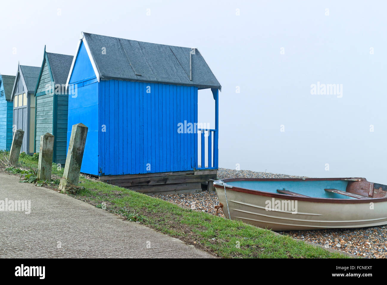 Blue green wooden beach huts and old fishing boat by a footpath in English Kentish seaside resort of Margate on a foggy day Stock Photo
