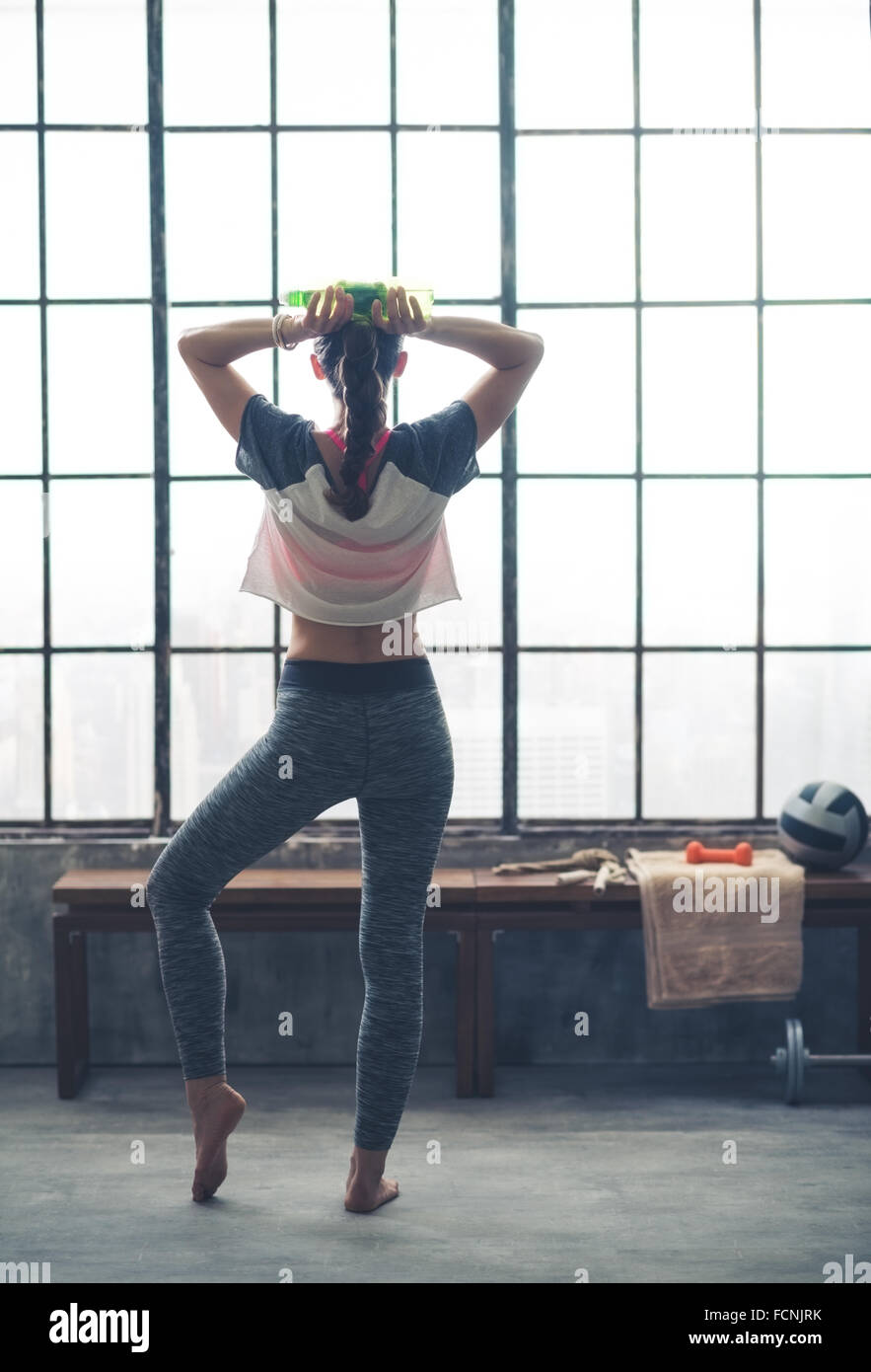 Seen from behind, a woman stands casually stretching out one foot as she holds a bottle of water, resting her hands behind her head. Stock Photo