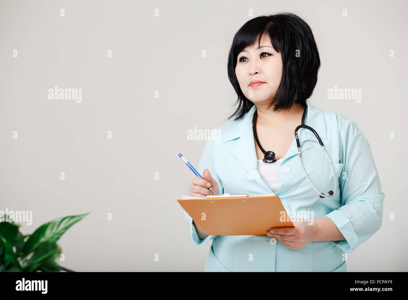 Curvy female nurse stands still writes the results by pen at paper, in medical lab coat with stethoscope around her neck. Asian appearance, dark short hair. Small vegetation on foreground. Stock Photo