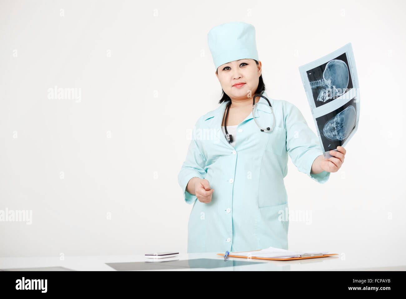 Curvy female doctor standing at table with X-ray examination results, image of skull in hand, wearing medical gown and cap, stethoscope. Asian, dark short hair. Radiology department. Stock Photo