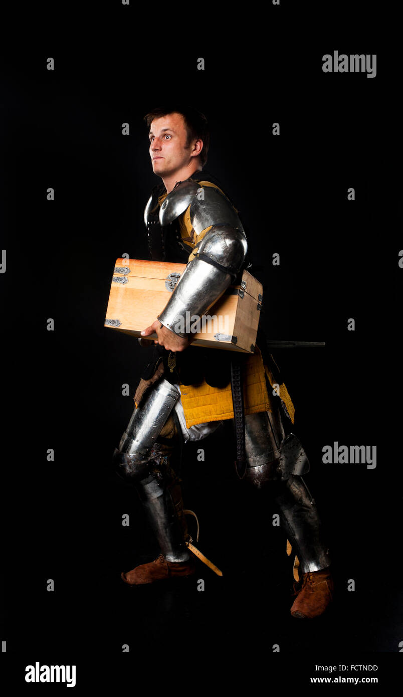 Humorous studio shot of young man dressed as medieval knight carrying away treasure chest (on black background) Stock Photo