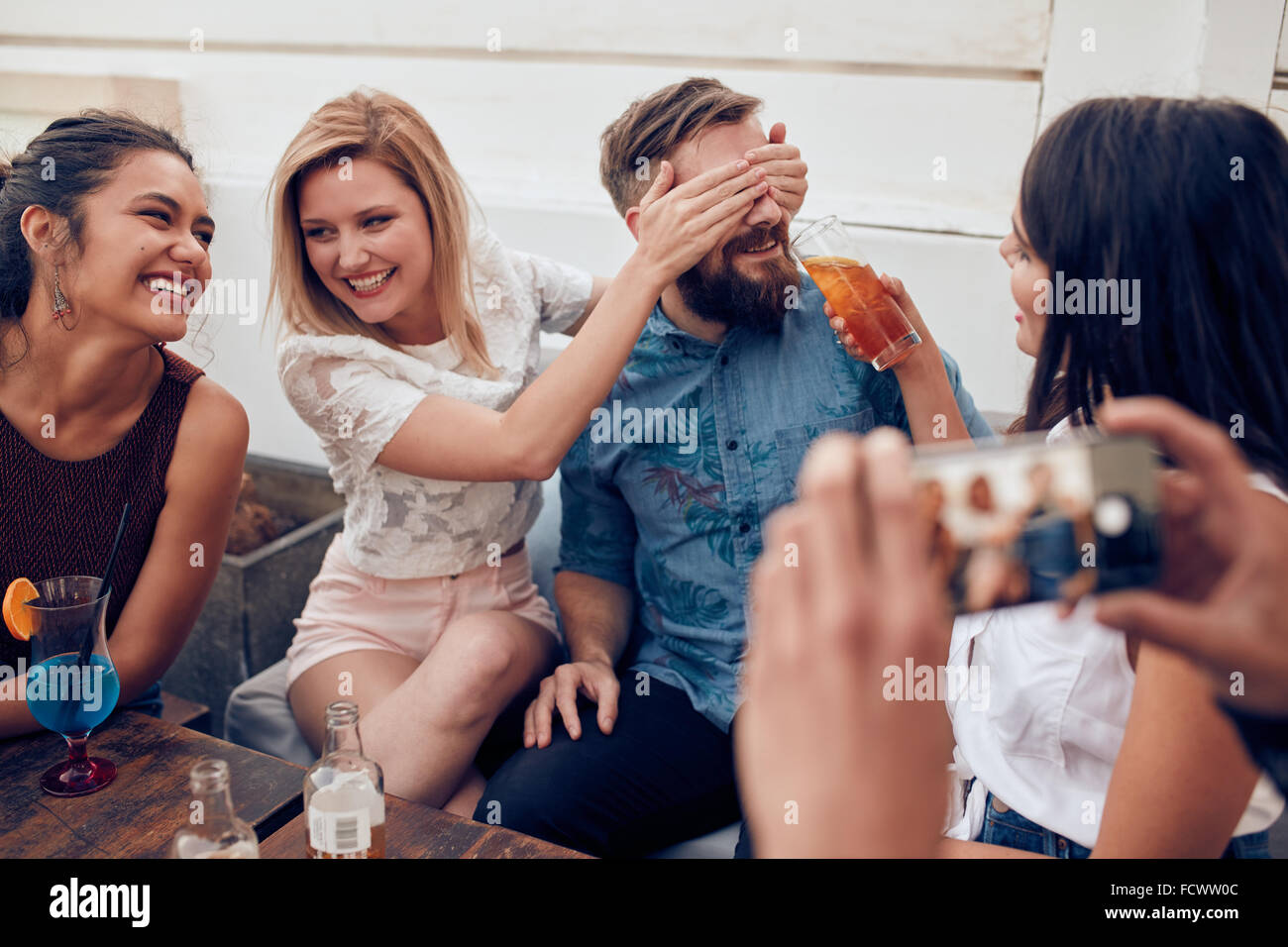 Young people sitting together enjoying party. Woman closing eyes of a man with another giving drink. Young friends having fun at Stock Photo
