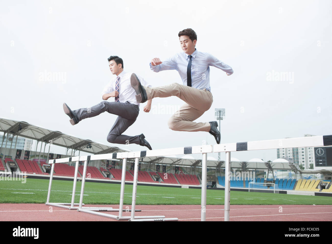 Two business men running and jumping over hurdles on a track with the background of sports field Stock Photo