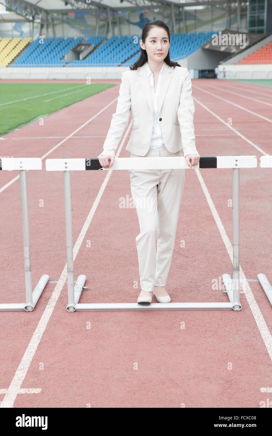 Woman in formal wear looking up and standing behind a hurdle on a track with the background of sports field Stock Photo