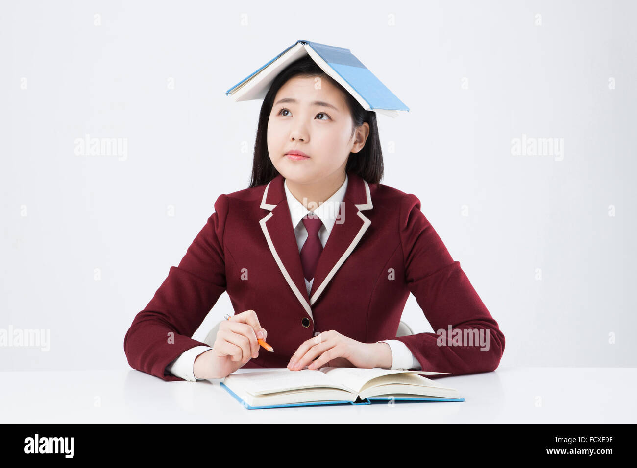 High school girl in school uniform seated at desk looking up with another book placed on her head Stock Photo