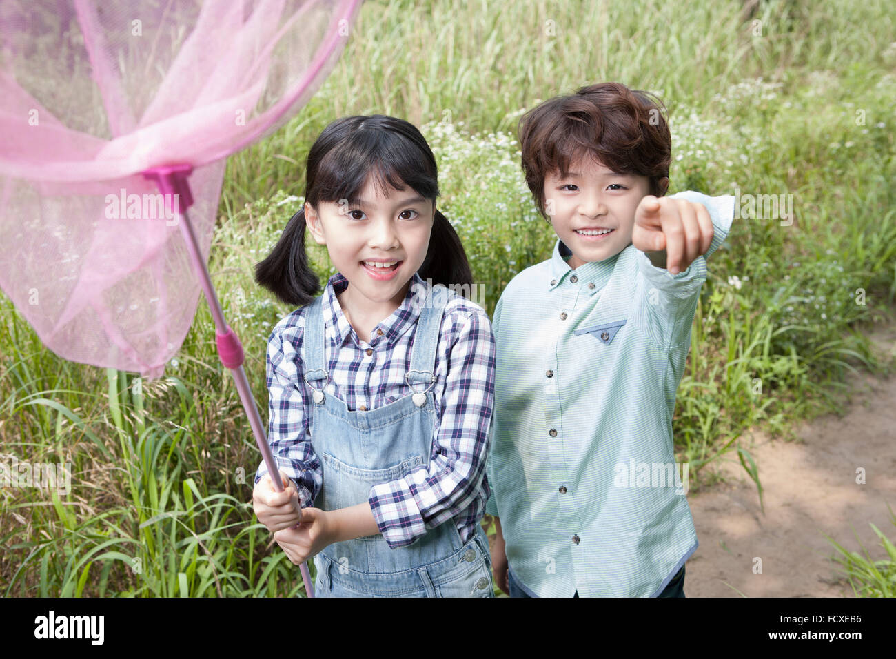 Two kids staring forward with a smile with a butterfly net at the grass field and the boy pointing with his finger forwards Stock Photo