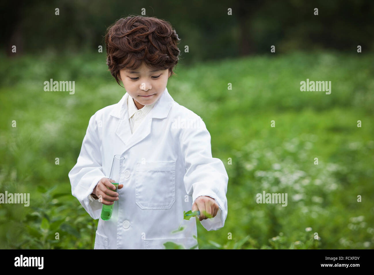 Boy in white gown holding a test tube and a leaf and looking down at field Stock Photo
