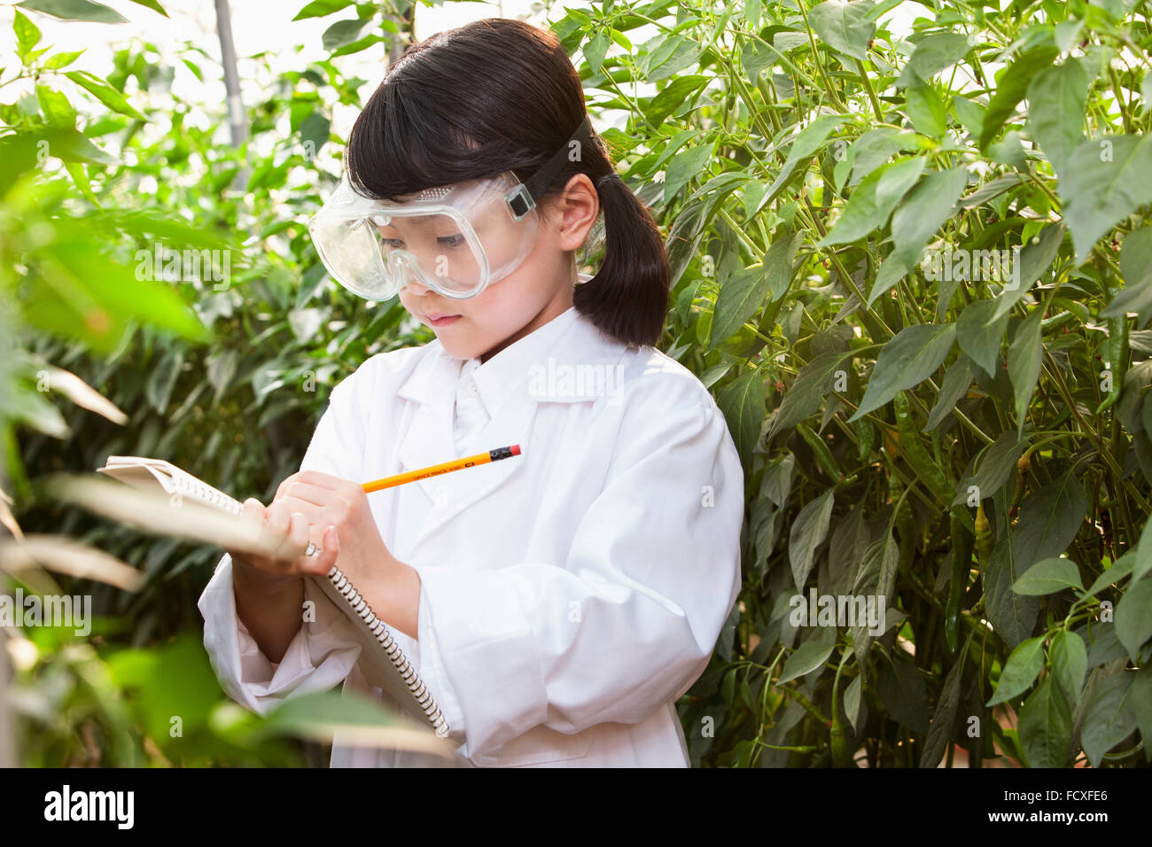 Girl in white gown and laboratory goggles writing down on a notebook surrounded by plants Stock Photo