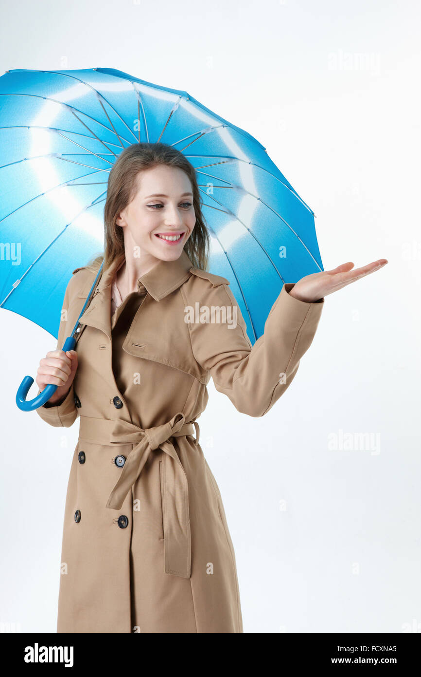 Young smiling woman with long hair wearng blue umbrella and trechcoat holding hand looking down Stock Photo