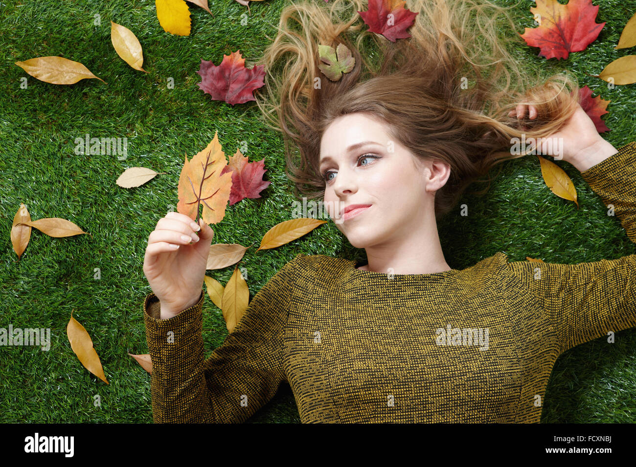 Portrait of young woman with long hair lying down on grass with fallen leaves Stock Photo