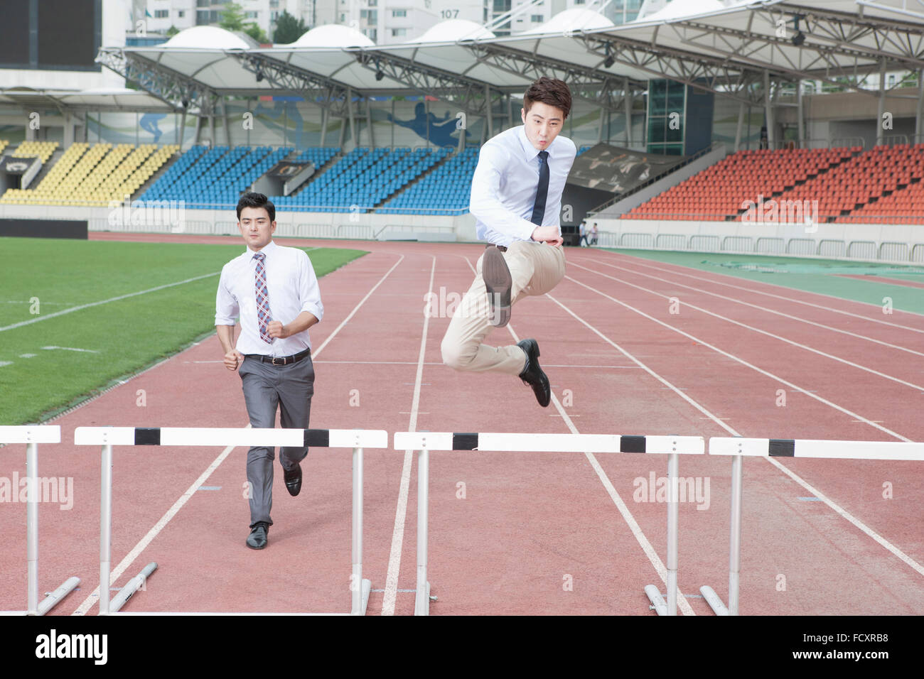 Businessmen running and jumping over hurdles on tracks at stadium Stock Photo