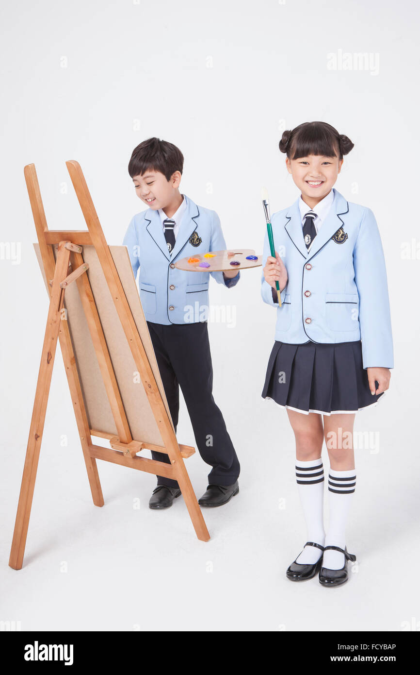 Elementary school girl standing with a brush and elementary school boy standing and drawing on easel holding a palette both in Stock Photo