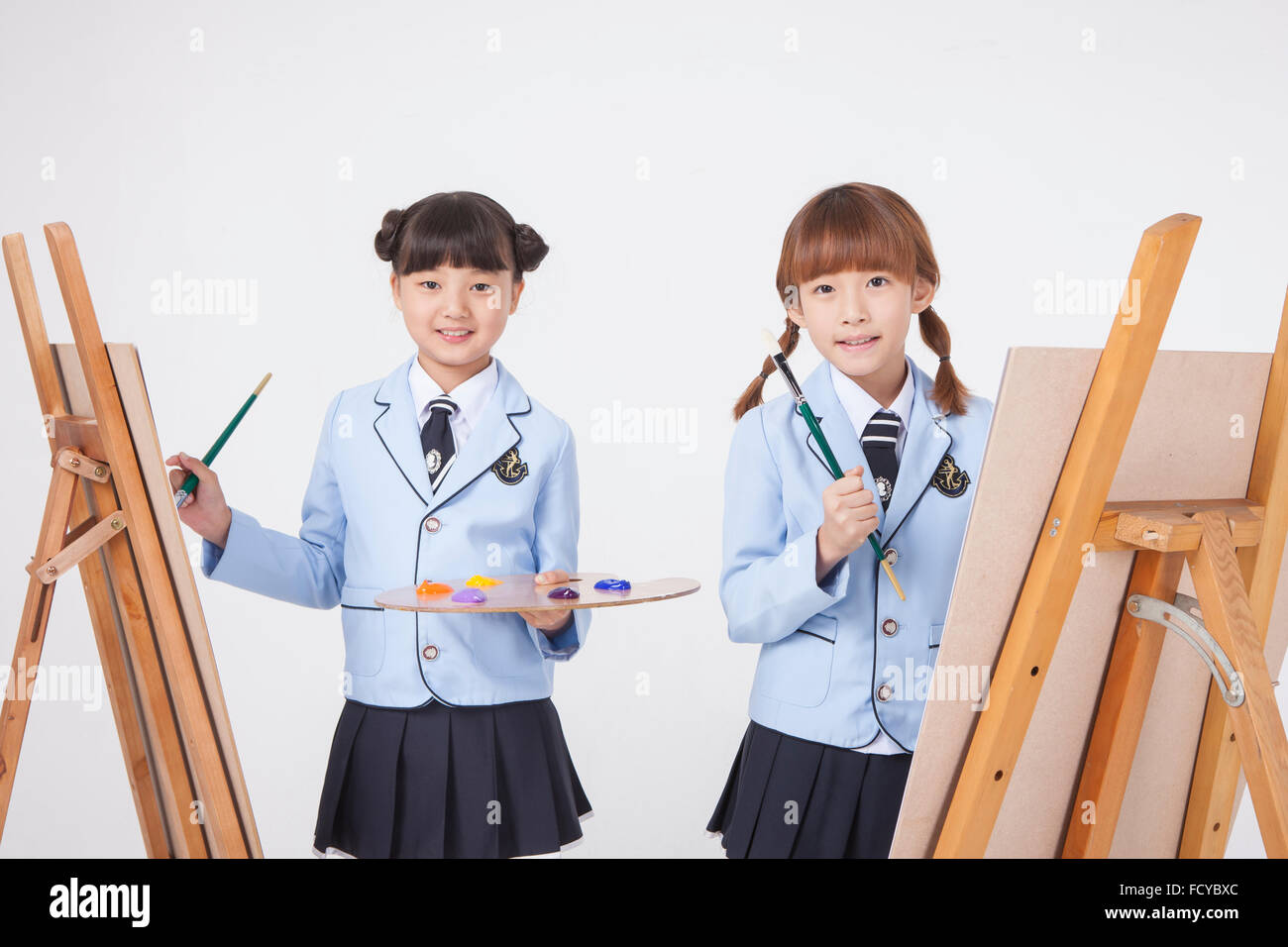 Two elementary school girl in art class holding a brush and a palette with an easel each both staring forward Stock Photo