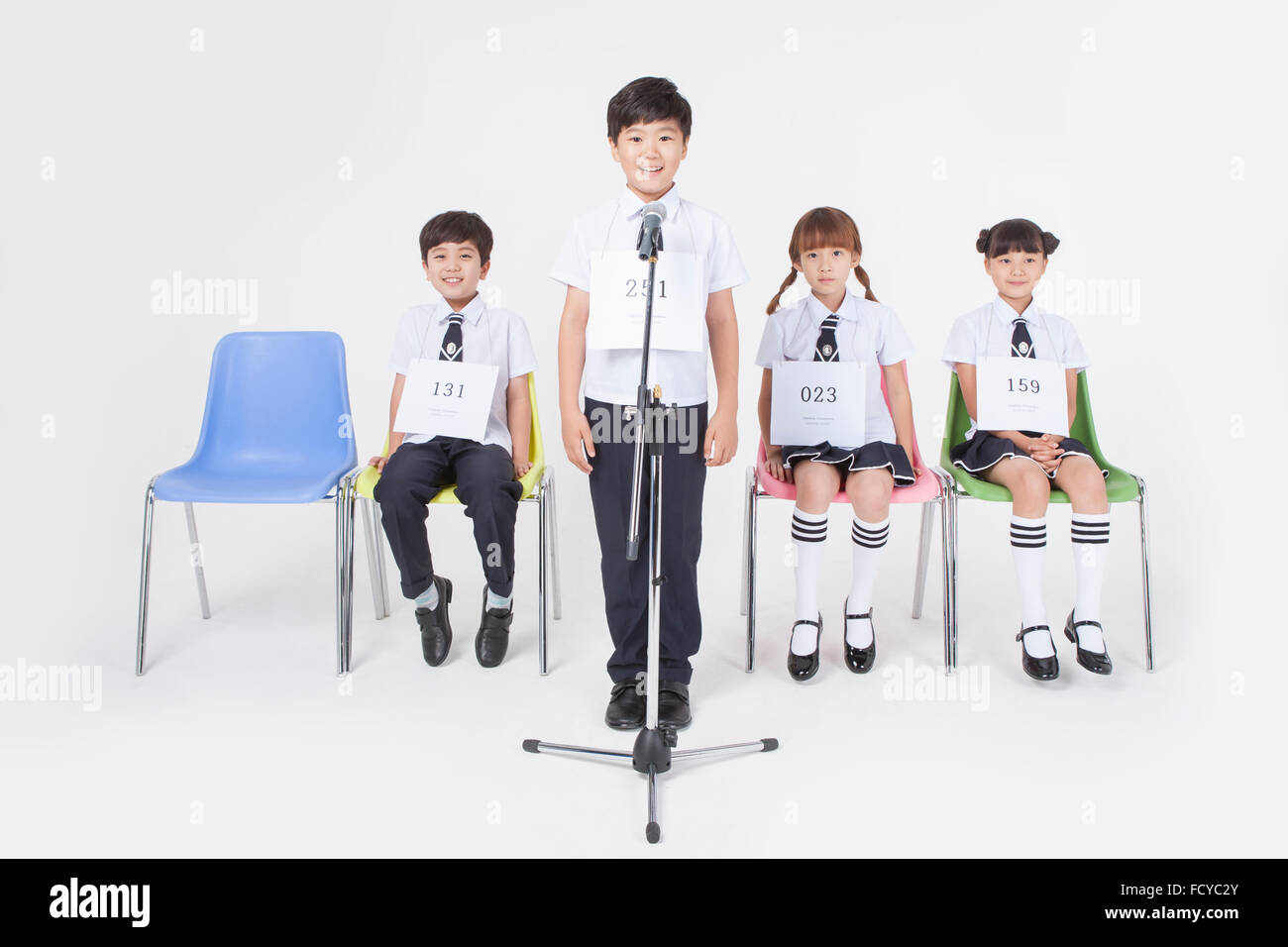 Elementary school boy standing behind a microphone and three other students seated on a chair behind him all in school uniforms Stock Photo