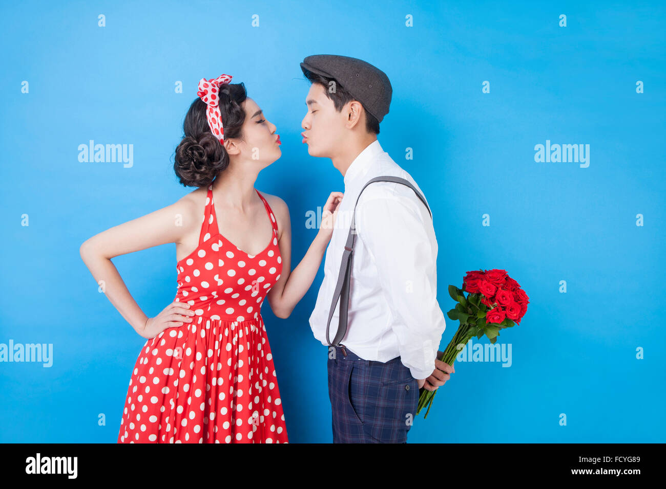 Couple in retro style fashion facing each other with the gesture of kissing and man holding roses behind his back Stock Photo